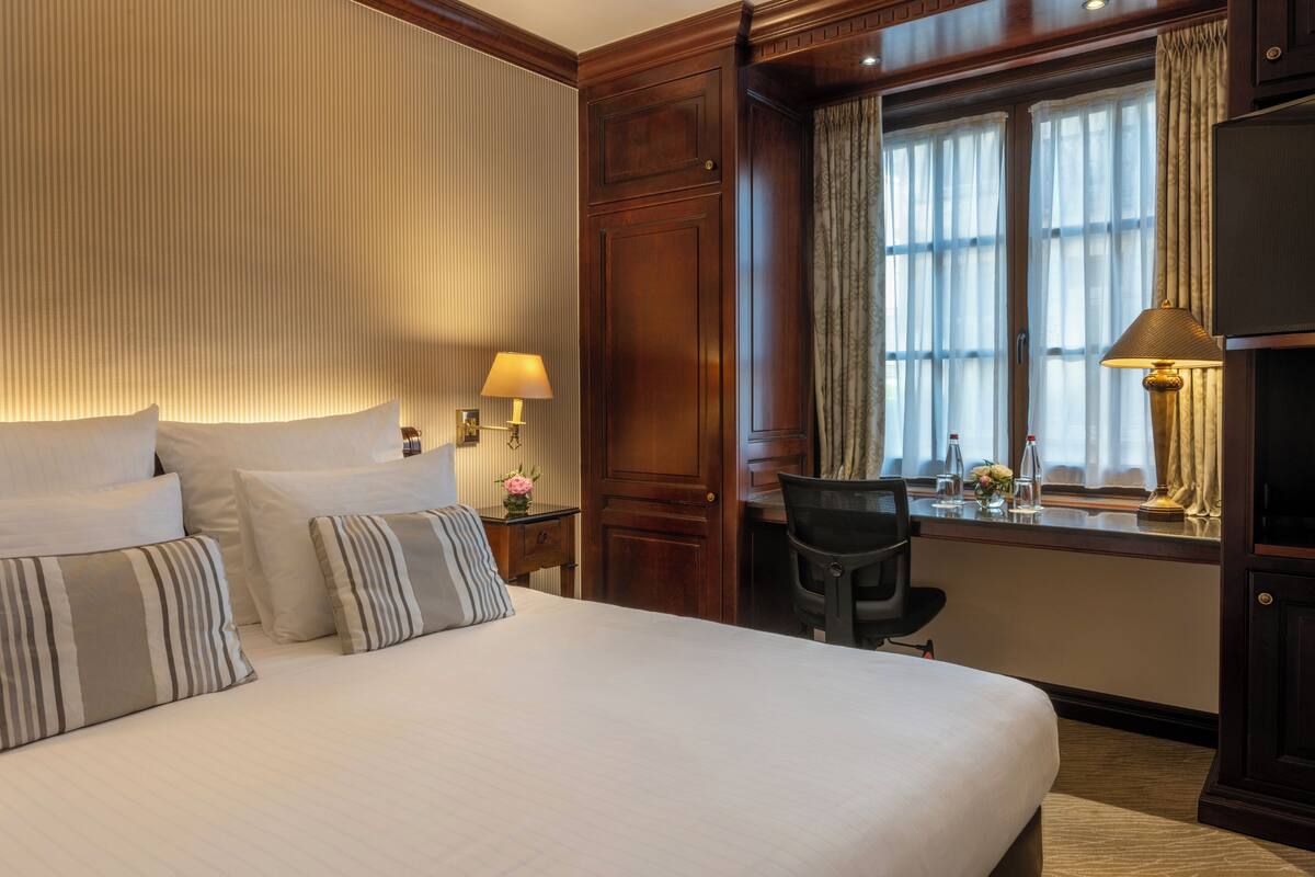 A hotel room with a large bed, a desk with a chair, a window with curtains, and a television.