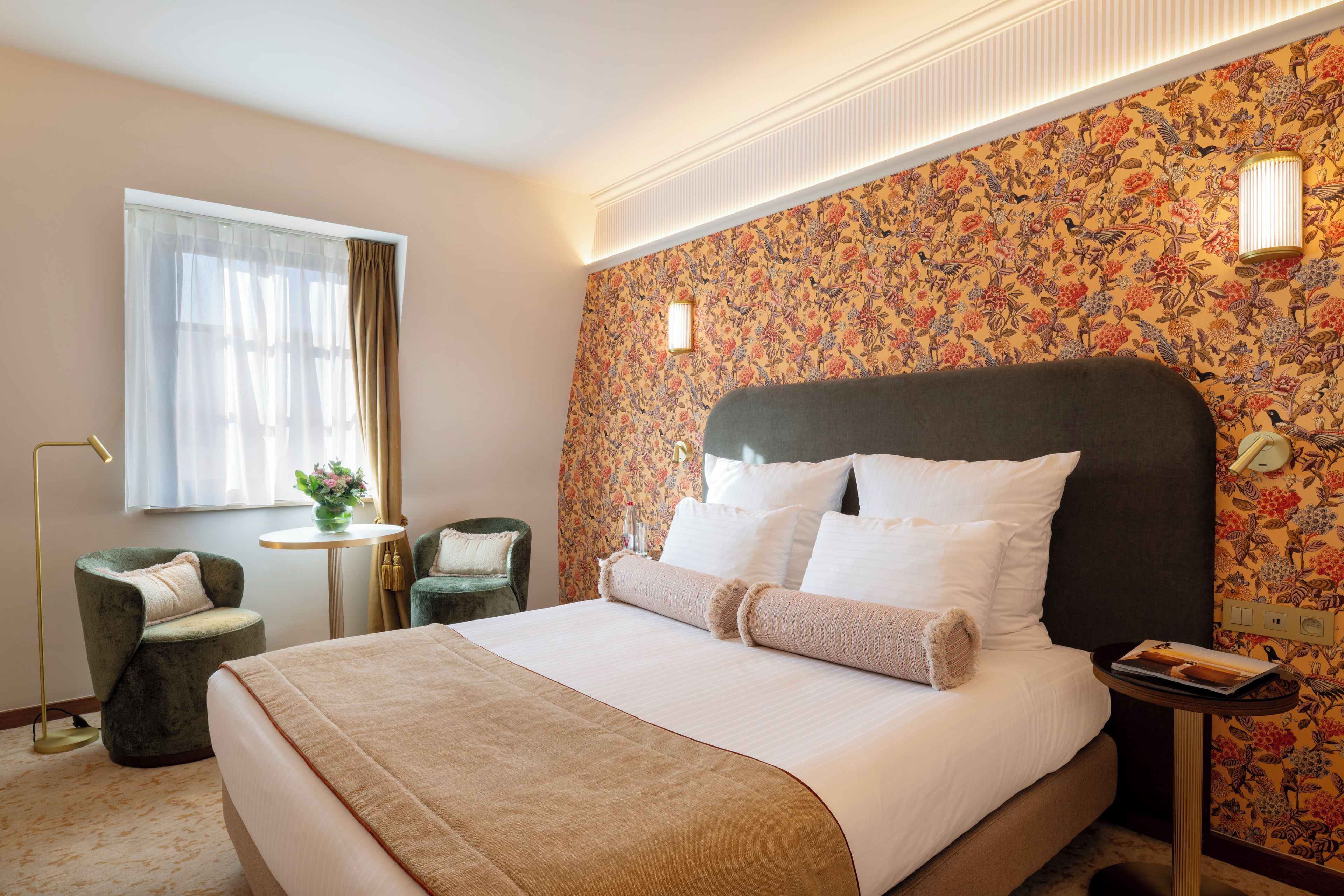 A hotel room with a large bed, a small table with a vase of flowers, two chairs, and a window with curtains.