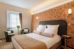 A hotel room with a large bed, a small table with a vase of flowers, two chairs, and a window with curtains.