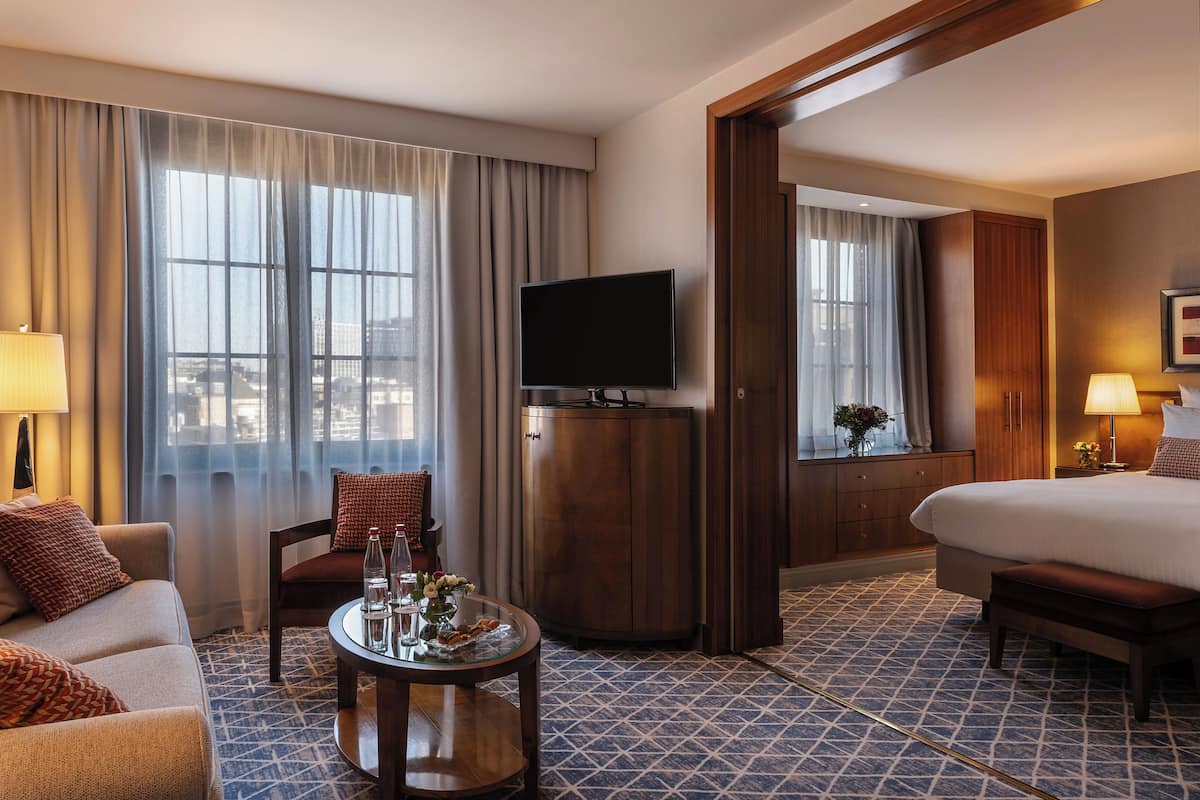 A hotel room with a bed, a TV, a desk with a lamp, a chair, and a view of the city through large windows.