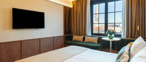 Una habitación de hotel con una cama grande, un televisor montado en la pared, una ventana con cortinas y una mesita con un florero.