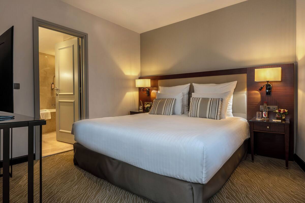 A hotel room with a large bed, two bedside lamps, a nightstand, a desk, and a shower area.