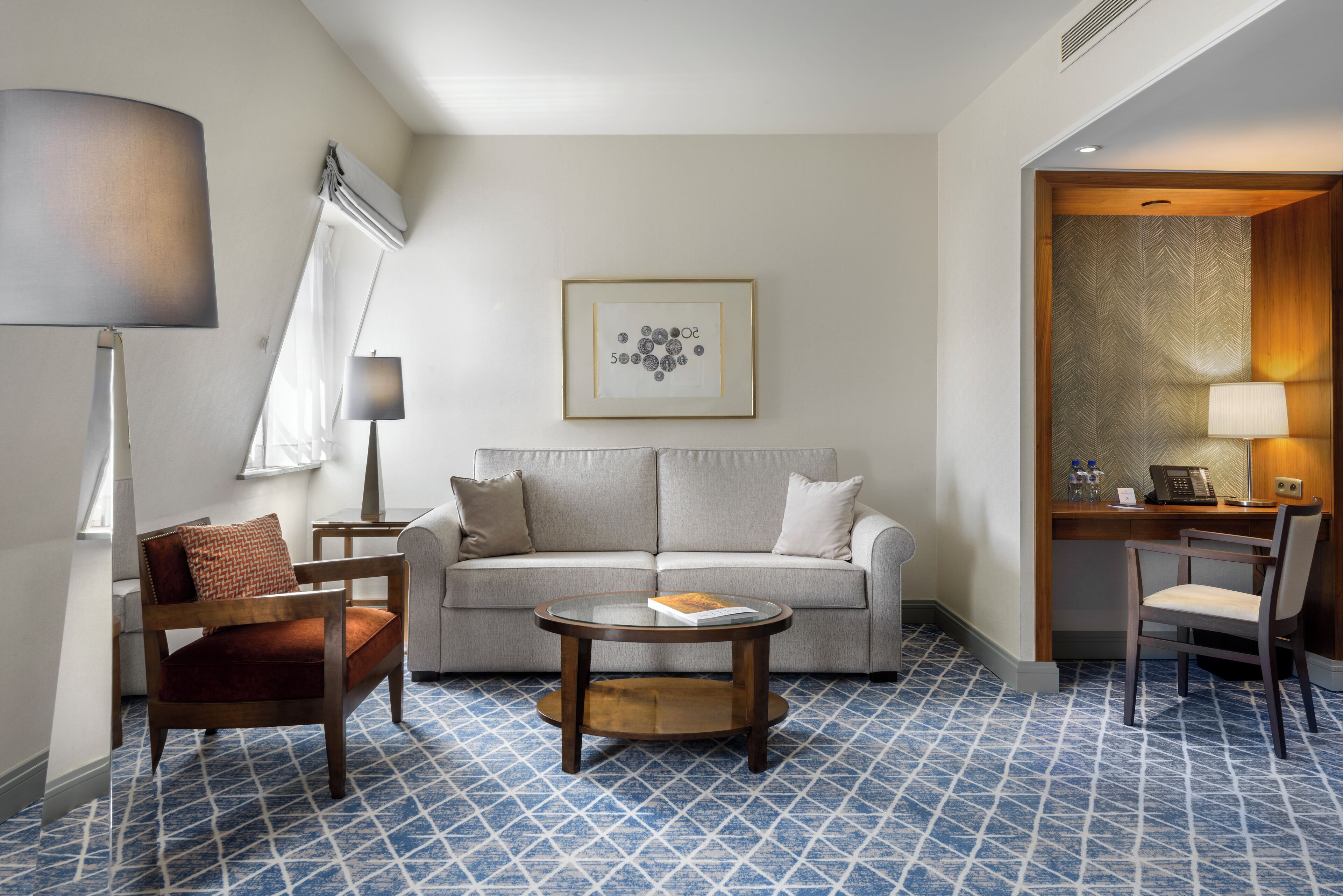 A hotel room with a sofa, a round coffee table, a wooden chair, a desk with a lamp, and a patterned floor.