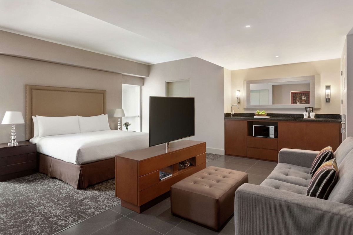 A hotel room with a bed, TV, sofa, ottoman, and kitchenette.