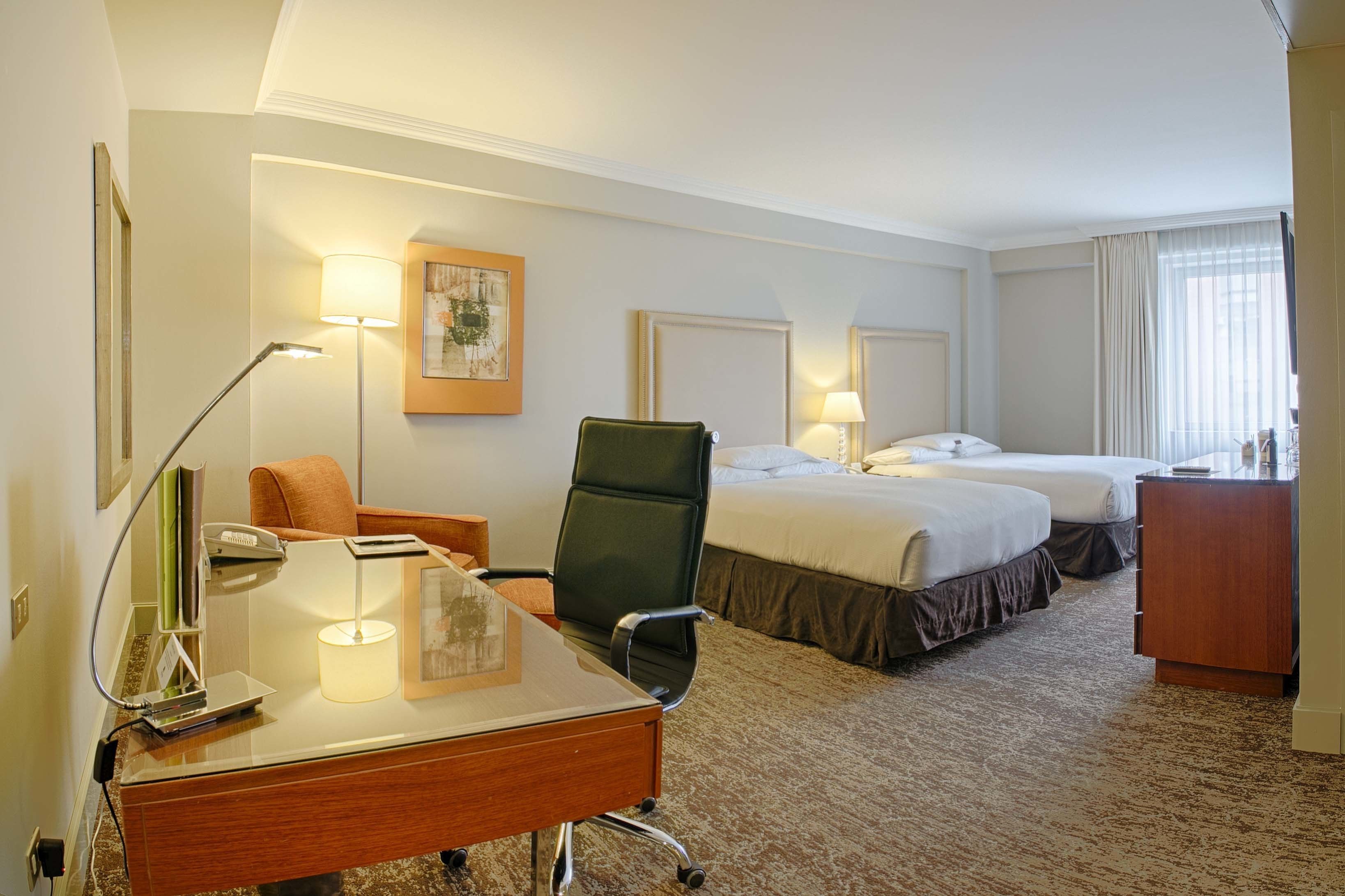 Una habitación de hotel con un escritorio de madera, dos camas, una silla y una lámpara.