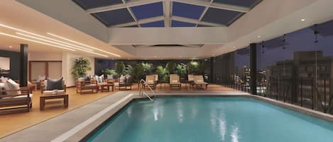 An indoor swimming pool with a glass ceiling, lounge chairs, and a view of the city at night.
