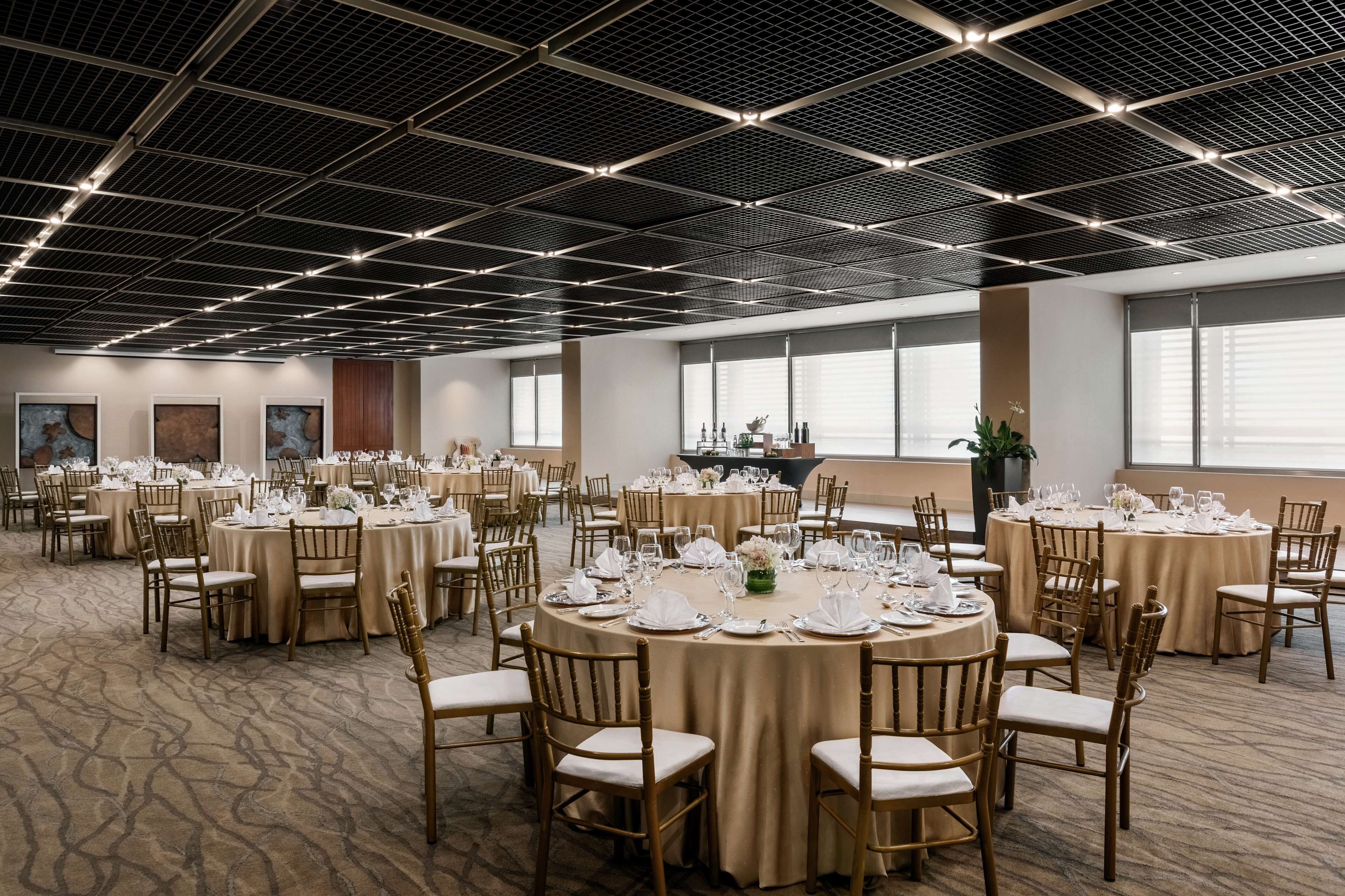 Un salón de banquetes con mesas redondas dispuestas para comer, con un diseño de techo moderno y grandes ventanales.