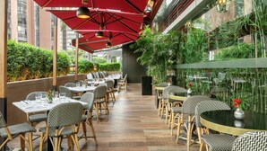 Un área de comedor al aire libre con mesas y sillas, sombrillas rojas y follaje.