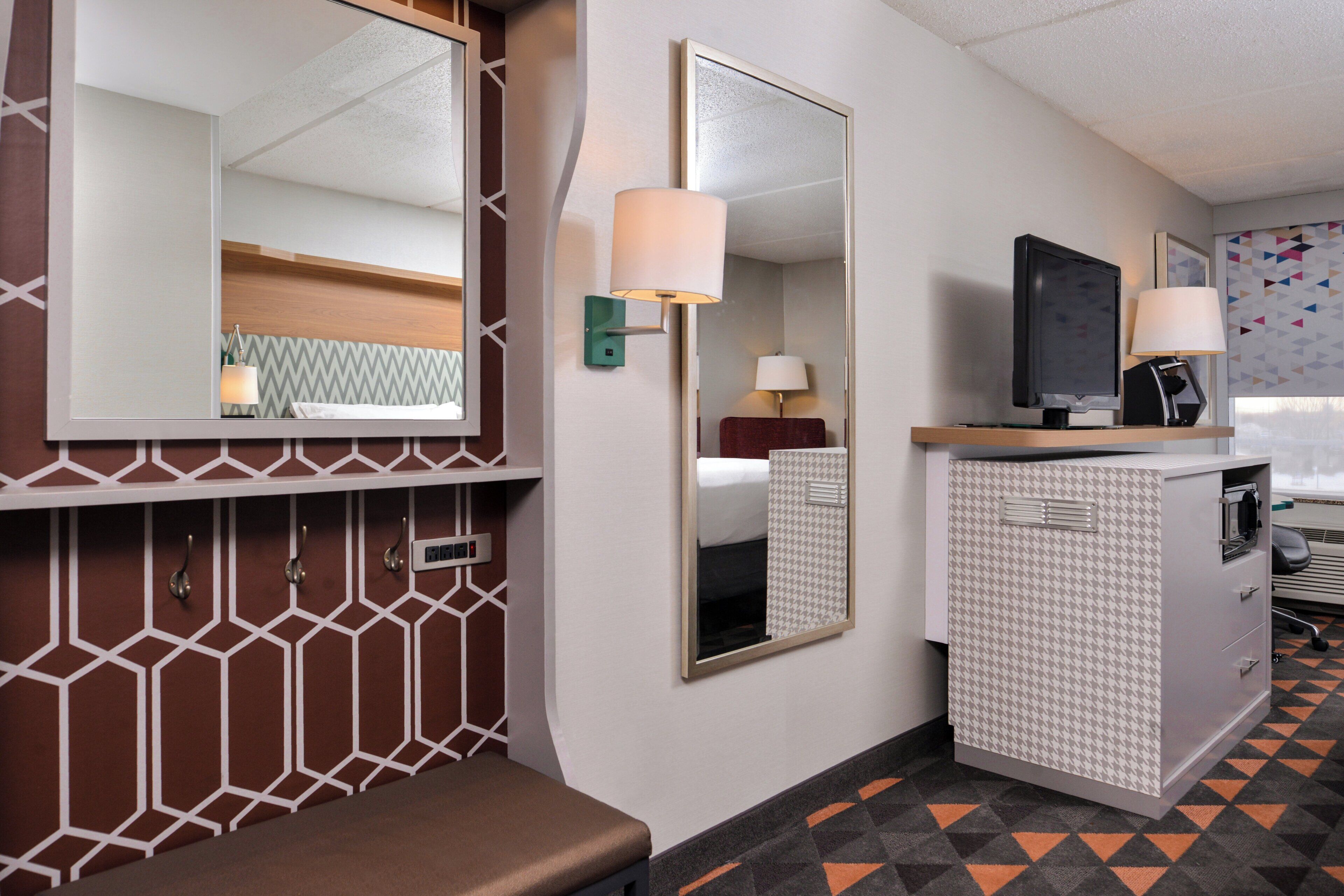 A hotel room with a bed, a desk with a computer, a mirror, and a small seating area.