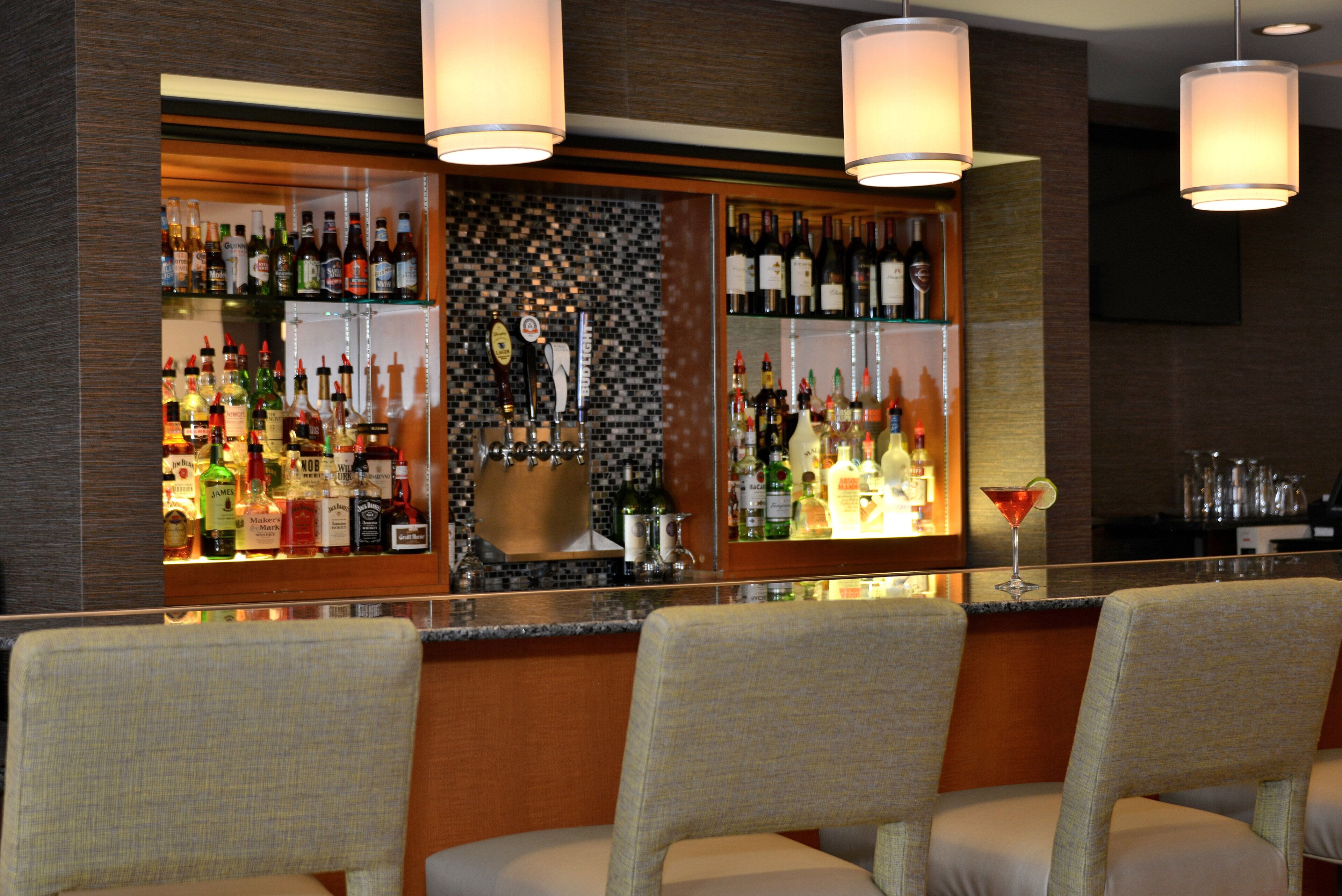 A bar area with multiple liquor bottles, a bar counter, and three upholstered bar chairs.