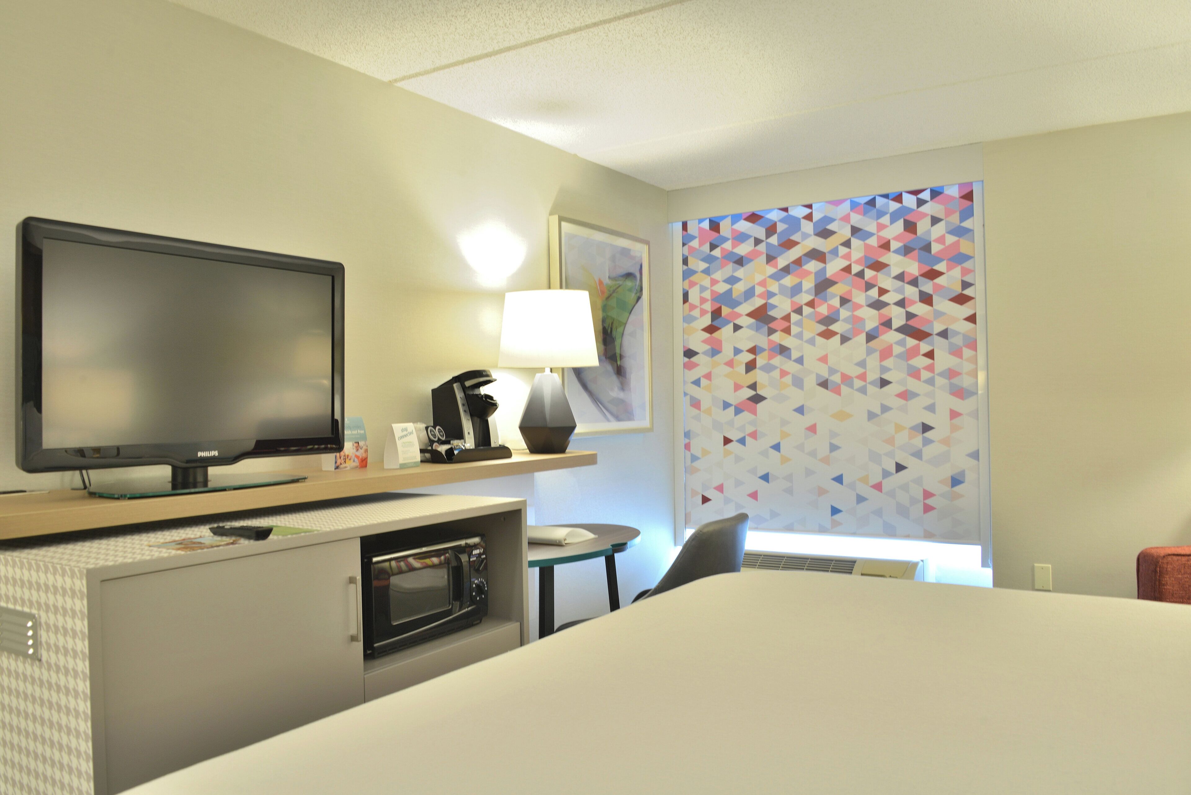 Habitación de hotel con televisor de pantalla plana, cafetera, lámpara, microondas, cama, escritorio y un colorido diseño geométrico en la pared.