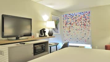 A hotel room with a flat-screen TV, a coffee maker, a lamp, a microwave, a bed, a desk, and a colorful geometric wall.