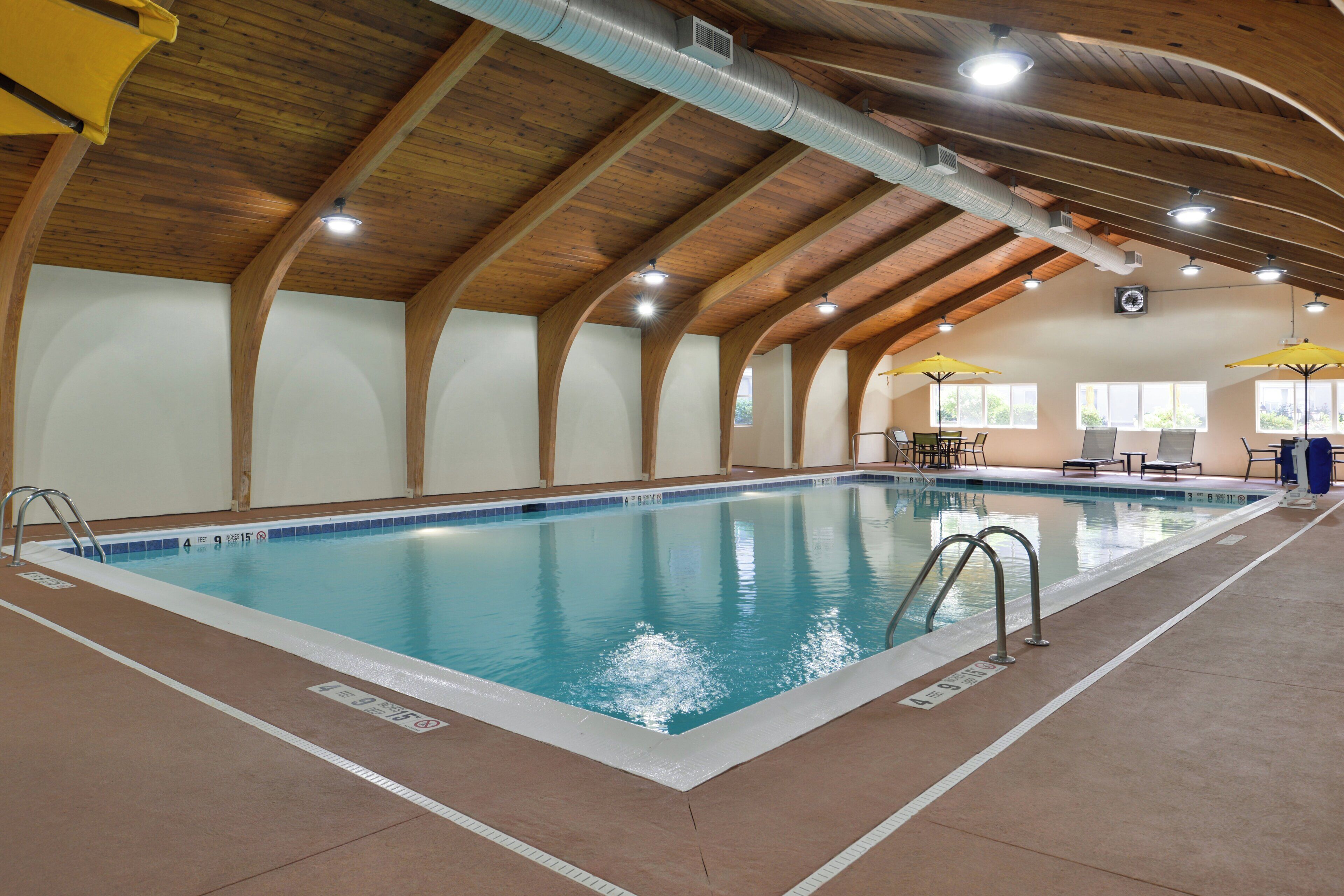 An indoor swimming pool with clear water, wooden beams on the ceiling, and a sunlit area with tables and chairs.