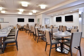 A conference room with multiple tables and chairs, equipped with water glasses and plates, and a flat-screen TV mounted on the wall.