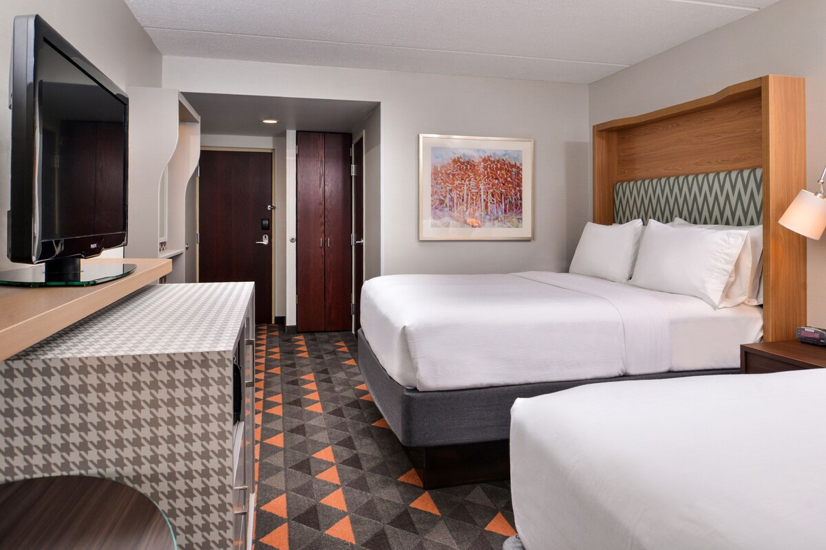 A hotel room with a bed, a TV, a desk, and a painting on the wall.