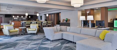 A modern hotel lobby with a sectional sofa, armchairs, a bar area, and a patterned carpet.