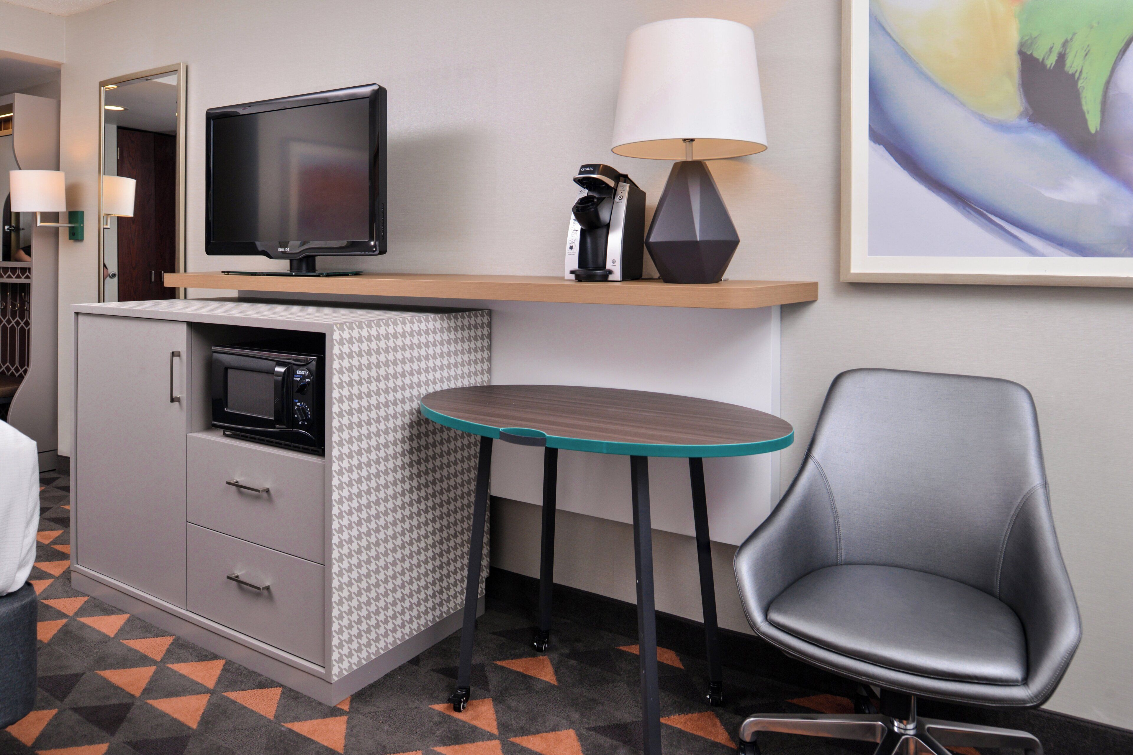 Habitación de hotel compacta con kitchenette, televisor de pantalla plana, lámpara, obra de arte enmarcada y una sola silla.