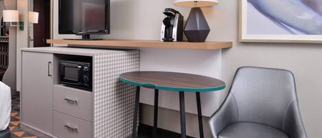 Habitación de hotel compacta con kitchenette, televisor de pantalla plana, lámpara, obra de arte enmarcada y una sola silla.
