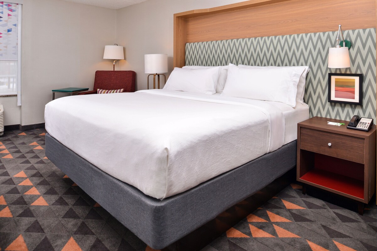 A hotel room with a large bed, a wooden headboard, a nightstand with a phone, and a wall-mounted lamp.
