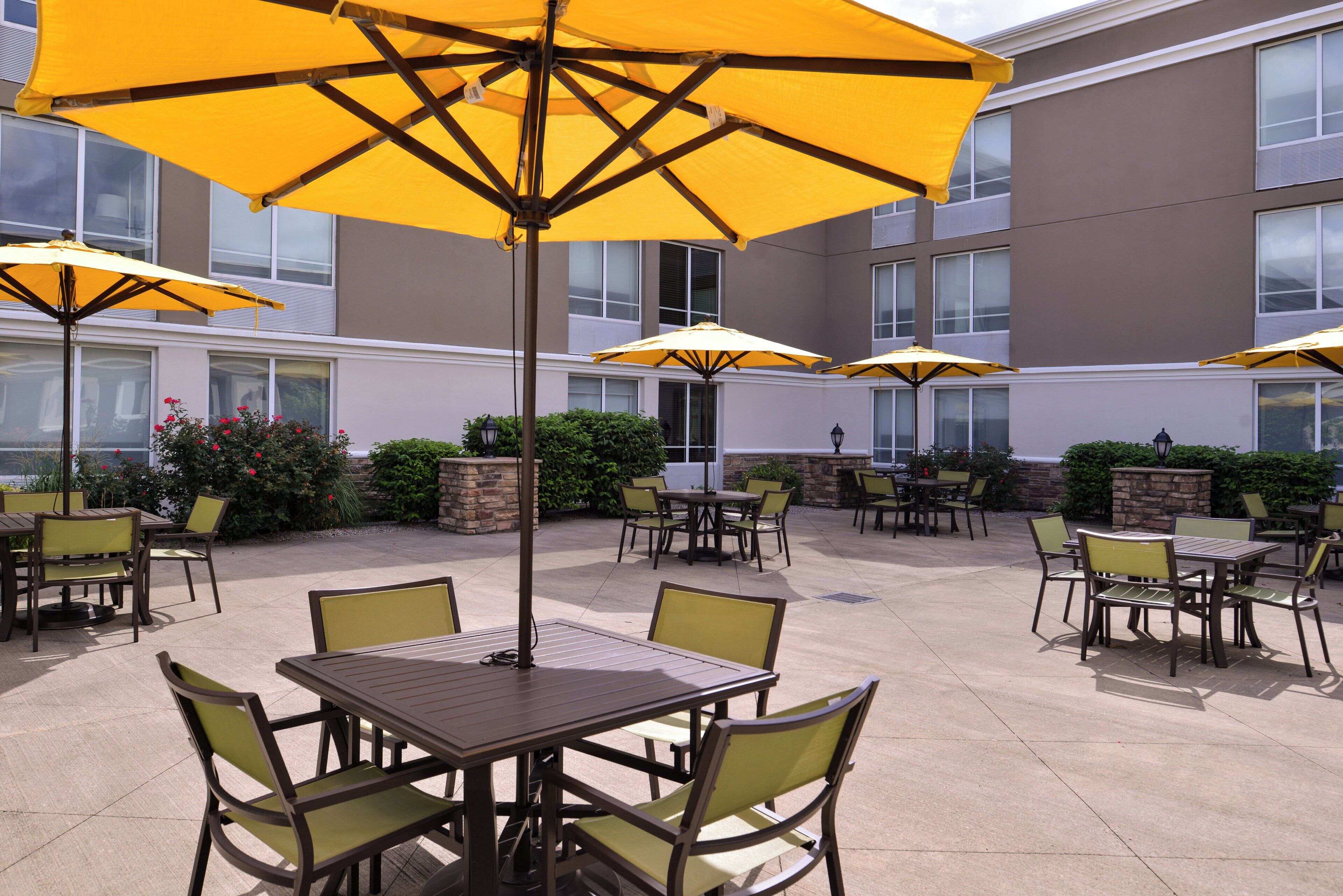 An outdoor patio area with multiple tables and chairs, some under yellow umbrellas, and a building with windows in the background.