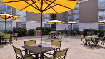 An outdoor patio area with multiple tables and chairs, some under yellow umbrellas, and a building with windows in the background.