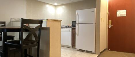 Una pequeña cocina americana equipada con refrigerador, microondas y lavavajillas. Hay una mesa de madera con sillas y una puerta a la derecha.