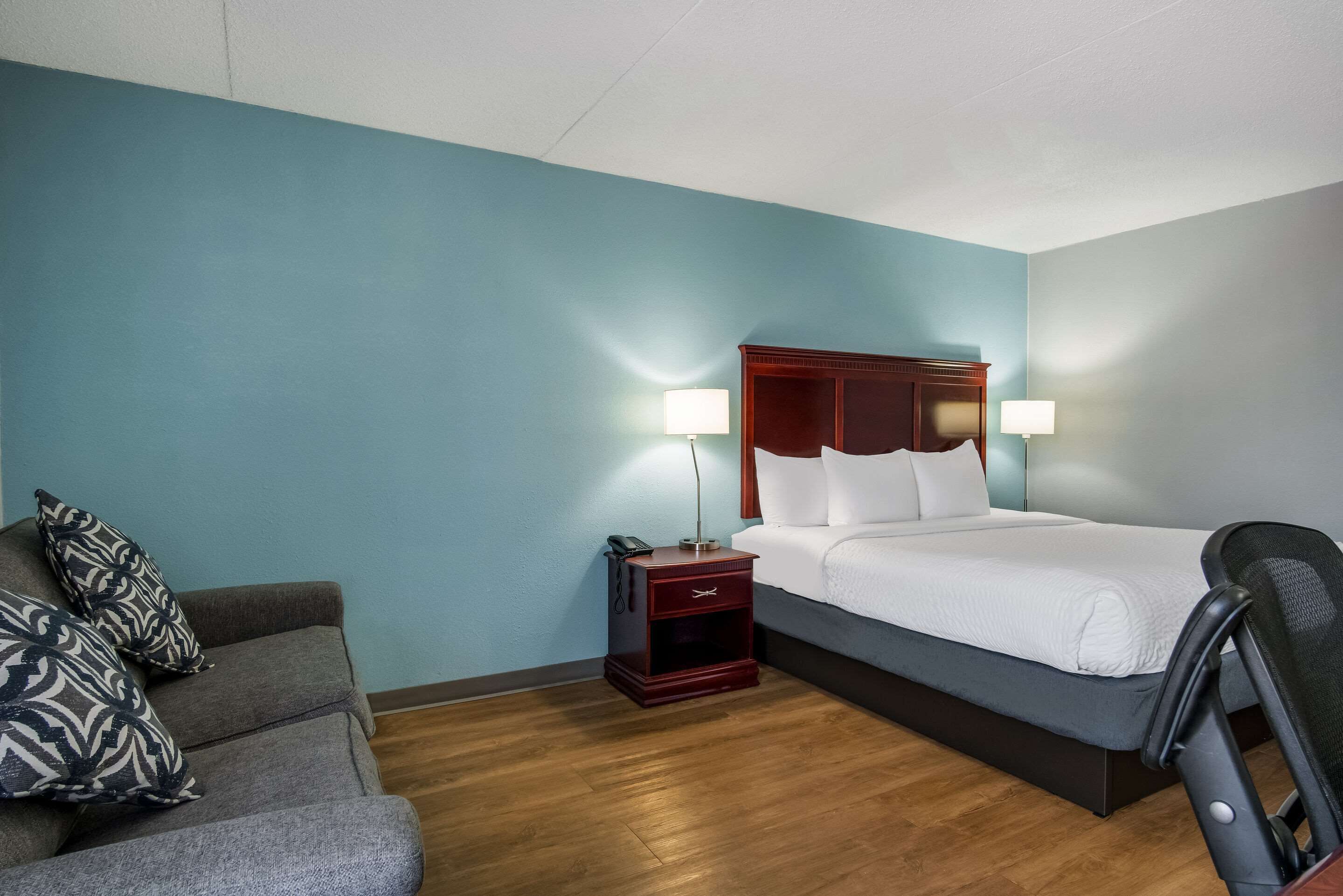 A hotel room with a bed, a nightstand with a lamp, a chair, and a sofa with patterned cushions.