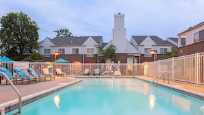 A swimming pool with lounge chairs and umbrellas, surrounded by a fence and buildings in the background.