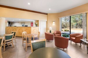 Un vestíbulo de hotel con mostrador de recepción, sillas de madera, una mesa redonda y grandes ventanales que ofrecen vistas al exterior.