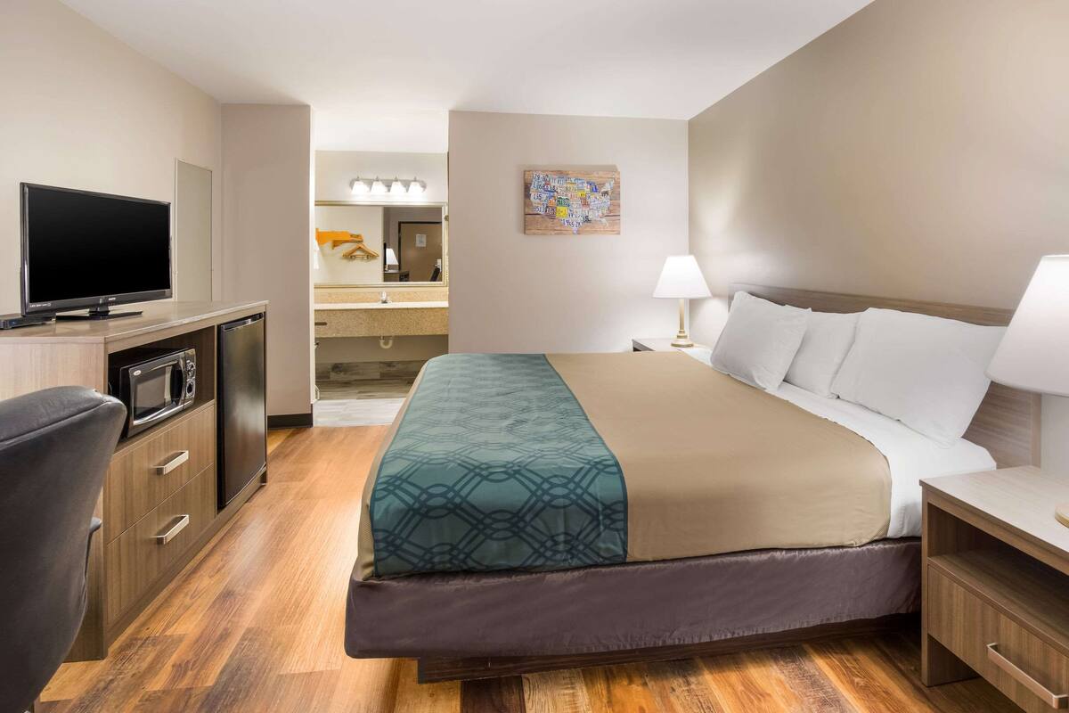 A hotel room with a bed, a desk, a television, a microwave, and a bathroom visible in the background.
