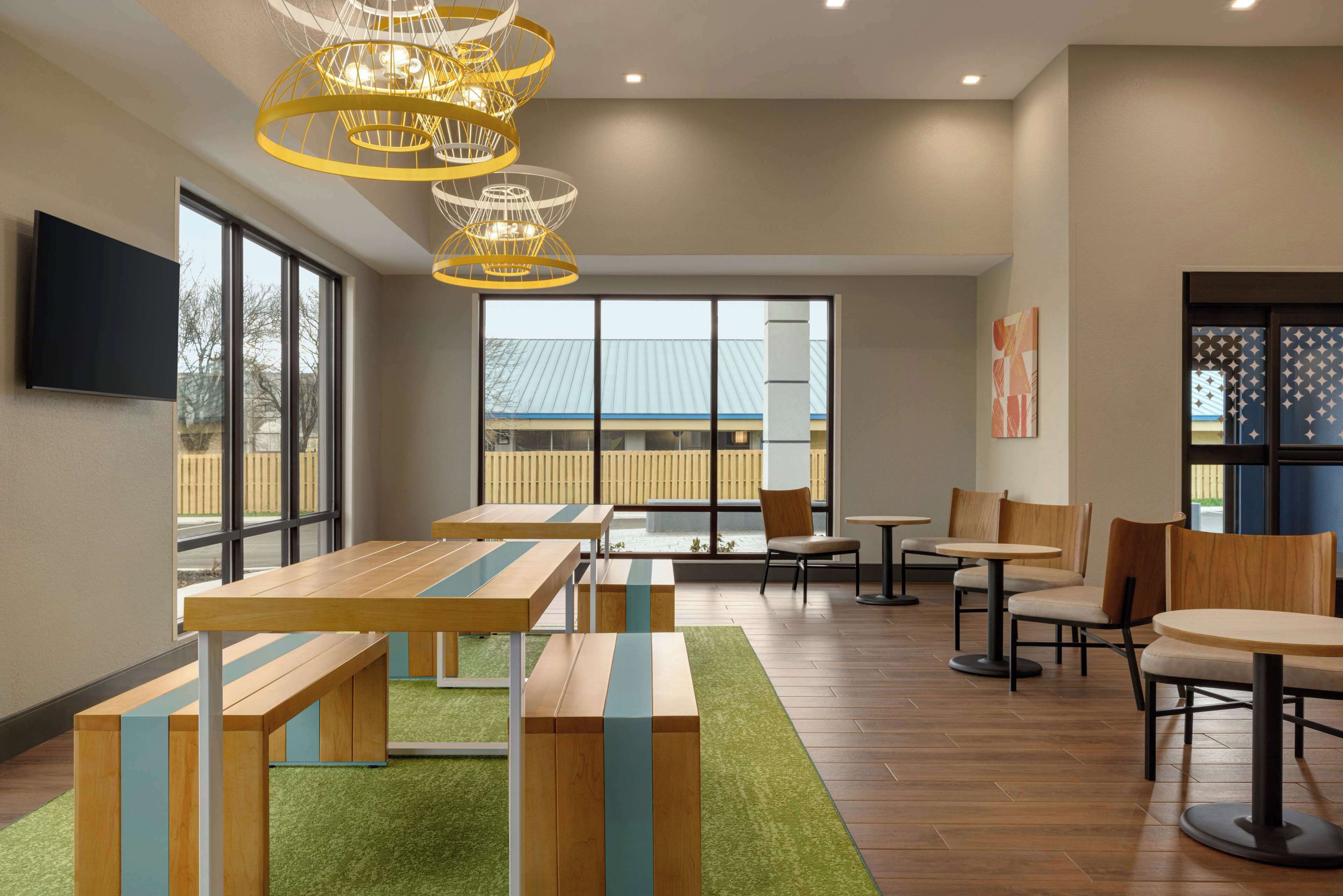 A modern indoor space with wooden tables and chairs, a green carpet, and large windows.