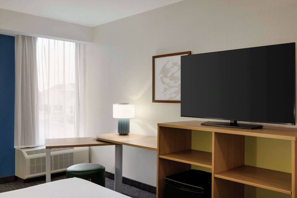 A hotel room with a bed, a desk with a lamp, a TV on a wooden stand, a radiator, and a window with curtains.