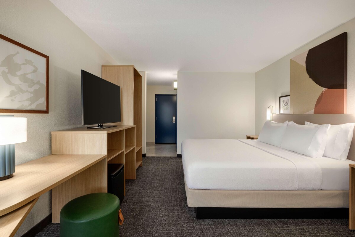 A hotel room with a bed, a desk, a TV, and a green stool.