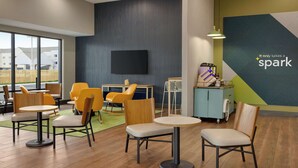 A modern indoor space with wooden flooring, yellow and beige chairs, and tables. A flat-screen TV is mounted on the wall, and there's a counter with various items. - Spark by Hilton Nashville at Opryland (Nashville)