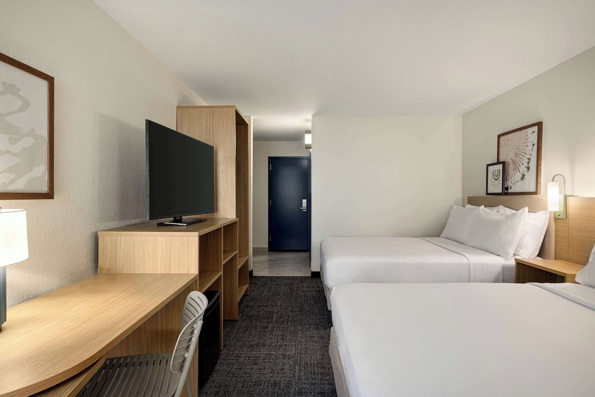 A hotel room with a desk, TV, two beds, and a door in the background.