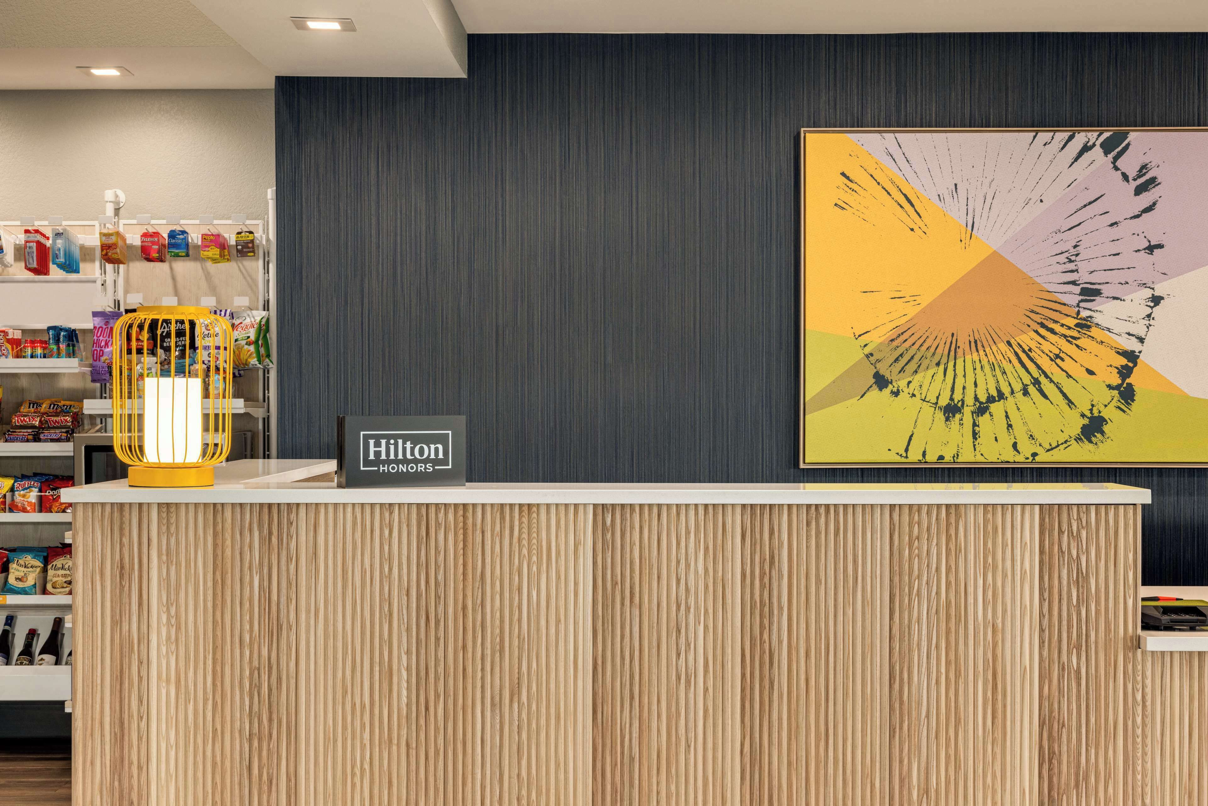 A hotel reception area with a wooden reception desk, a sign reading "Hilton HOMES," and a colorful abstract painting on the wall.
