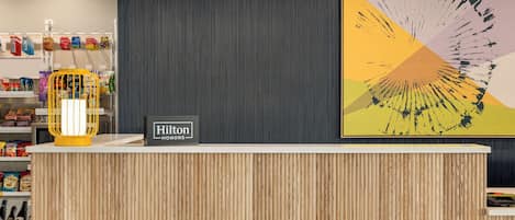 A hotel reception area with a wooden reception desk, a sign reading "Hilton HOMES," and a colorful abstract painting on the wall.