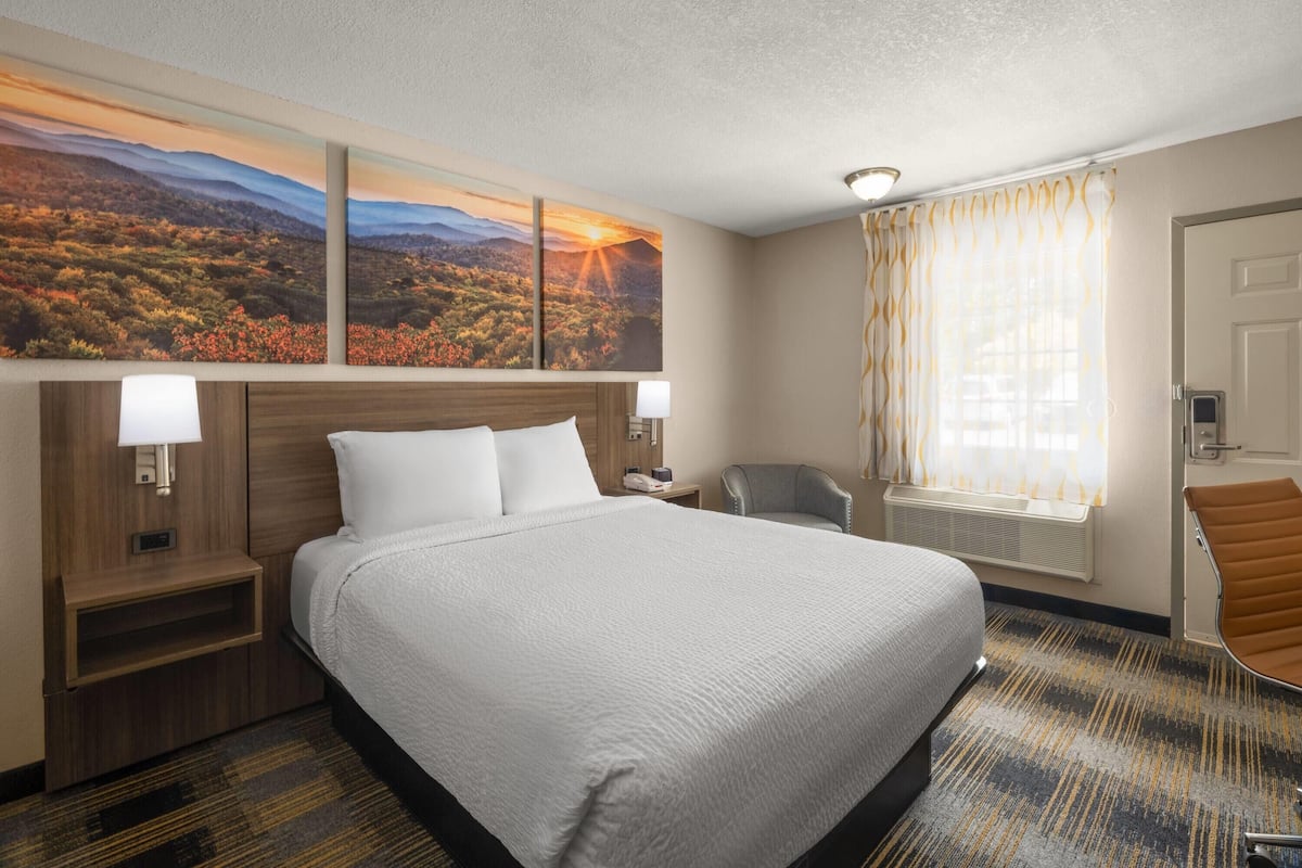 A hotel room with a large bed, a desk, a chair, a window with curtains, and a painting on the wall.