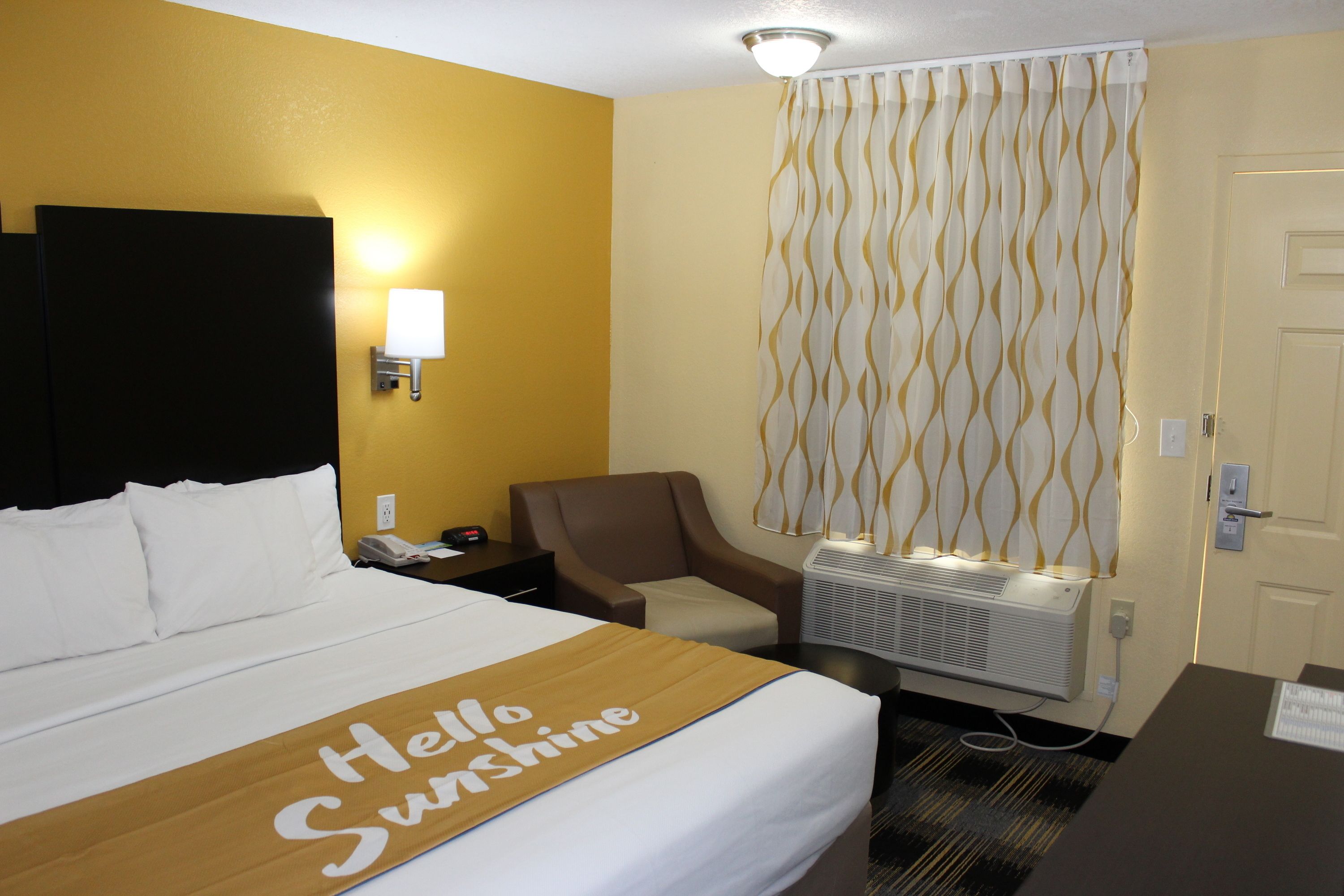 A hotel room with a bed, a chair, a desk, and a wall-mounted air conditioner.