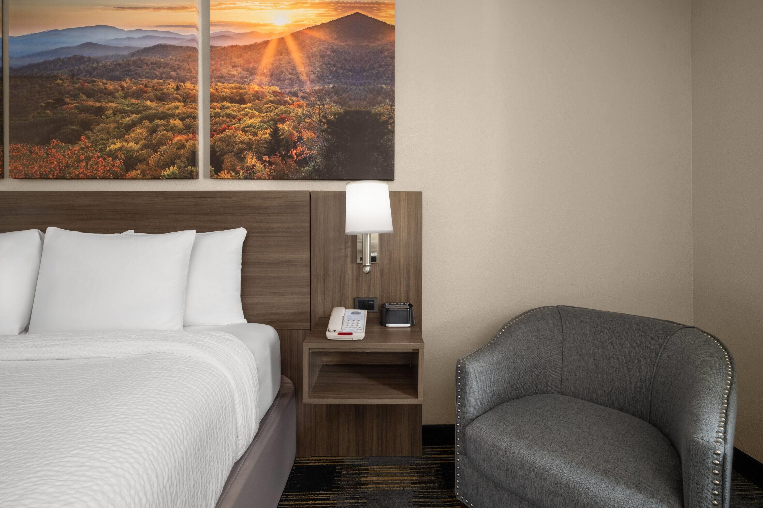 Quarto de hotel com cama, uma cadeira, mesa de cabeceira com luminária e um quadro de uma paisagem de pôr do sol.