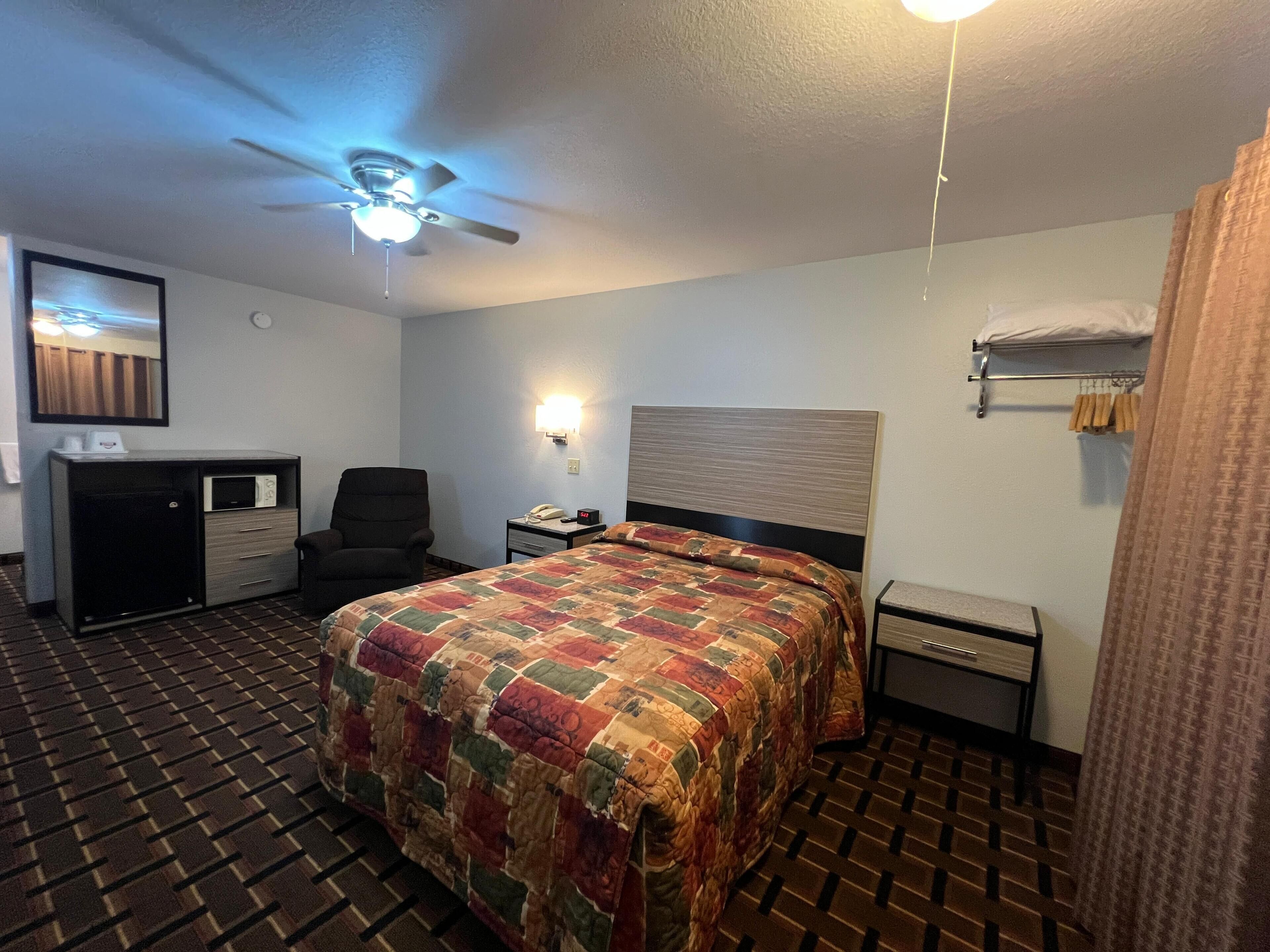 A hotel room with a bed, a desk, a chair, a ceiling fan, a nightstand, a lamp, a microwave, and a wardrobe.