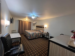 Hotellihuone, jossa on sänky, työpöytä, tuoli, kattotuuletin ja pieni pöytä, jonka päällä on maljakko täynnä kukkia.