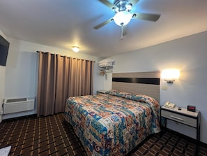 Hotellihuone, jossa on sänky, yöpöytä, jossa on puhelin ja kello, kattotuuletin, seinään kiinnitetty valaisin ja ikkuna, jossa on verhot.