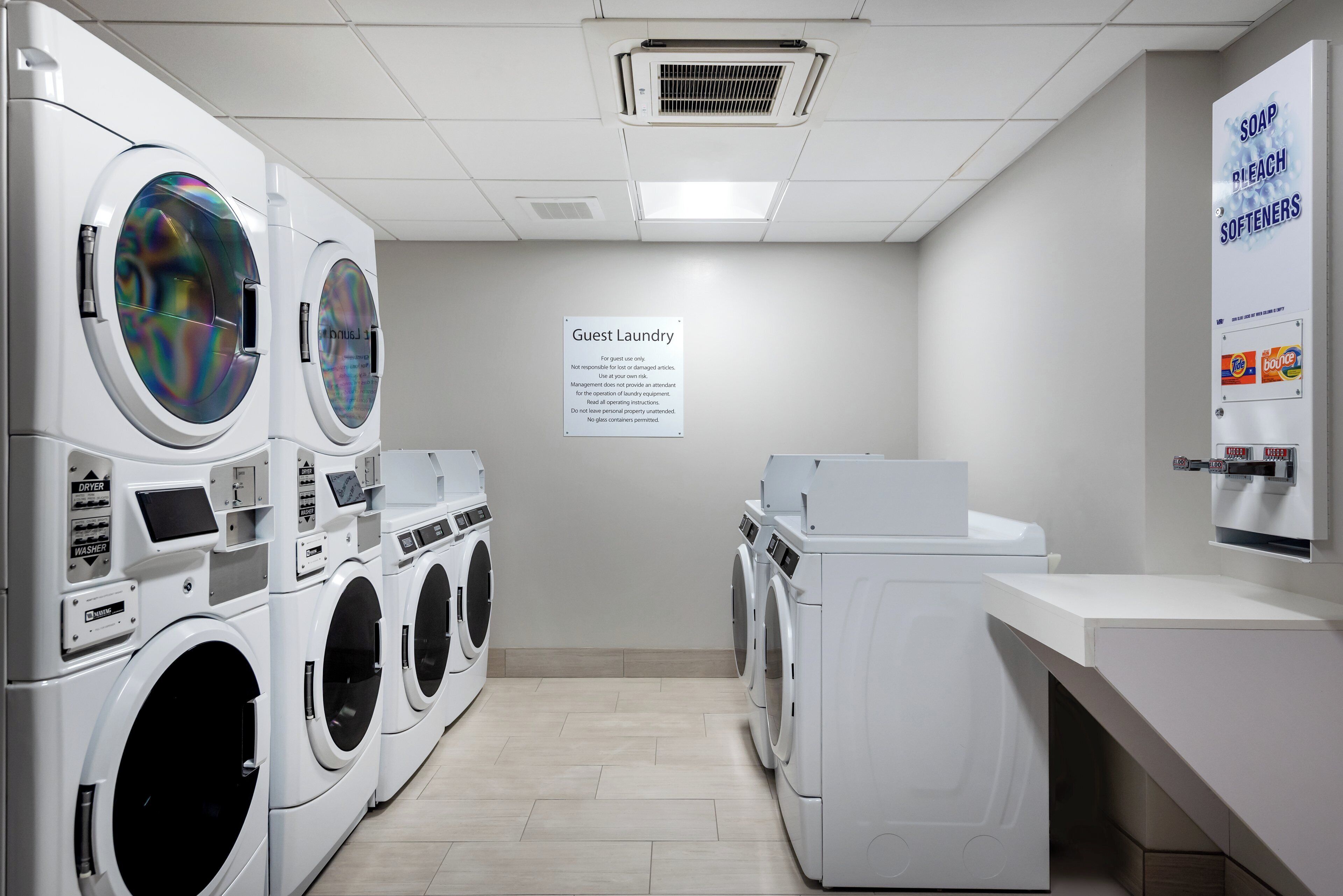 Une buanderie équipée de plusieurs machines à laver et de plusieurs sèche-linge, un panneau indiquant qu’elle est réservée aux clients, et un distributeur de savon et d’eau de Javel fixé au mur.