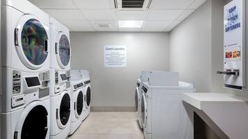Une buanderie équipée de plusieurs machines à laver et de plusieurs sèche-linge, un panneau indiquant qu’elle est réservée aux clients, et un distributeur de savon et d’eau de Javel fixé au mur.