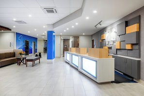 A modern reception area with a reception desk, seating area, and a counter with a digital display. - Holiday Inn Express & Suites Santa Ana - Orange County by IHG (Santa Ana)