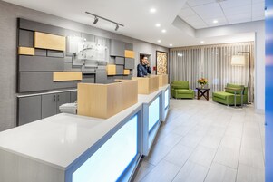A modern hotel lobby with a reception desk, green seating area, and a large window with curtains.