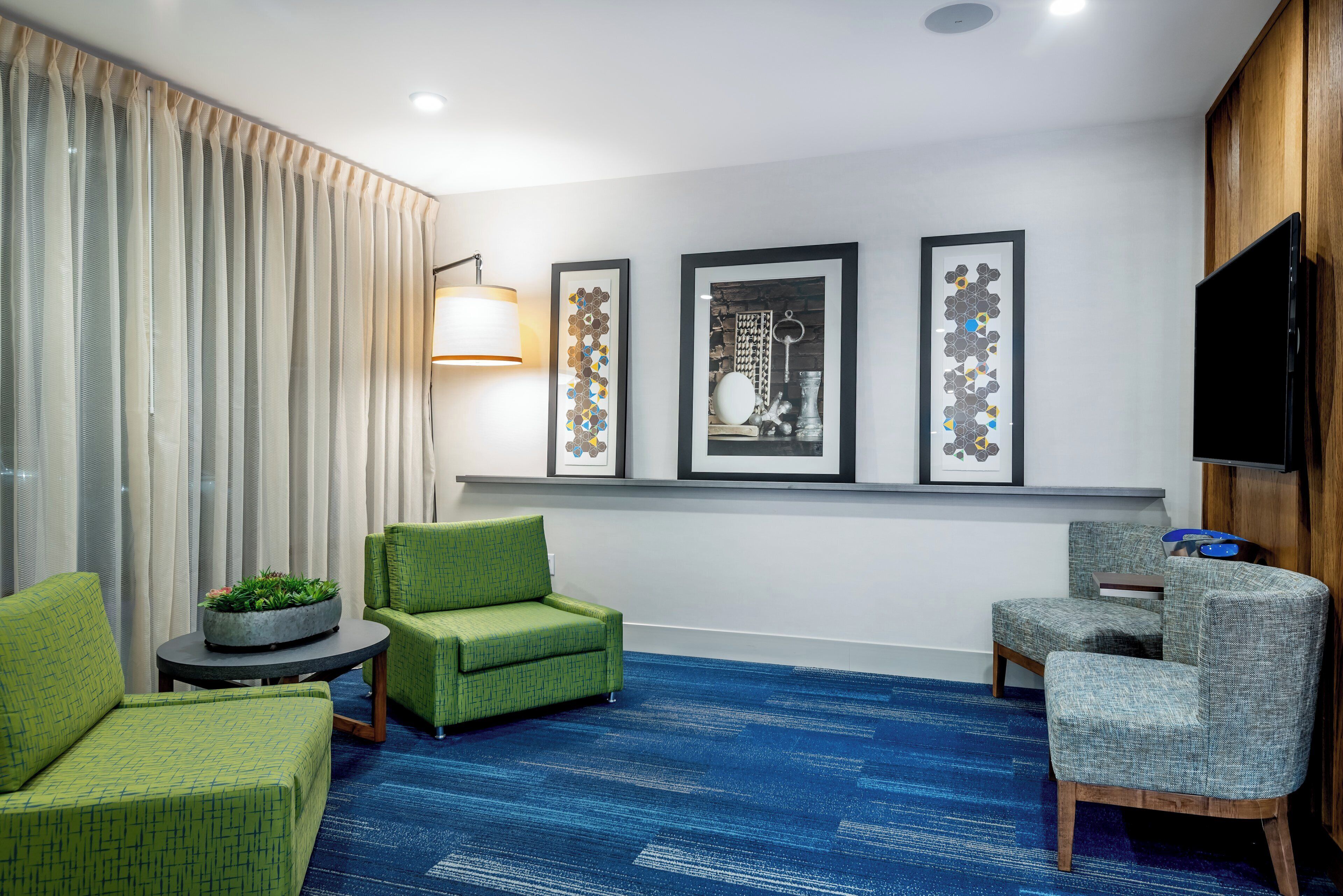 Una camera d'hotel con tappeto blu, poltrone rivestite in verde e grigio, un tavolino rotondo, una pianta in vaso e tre quadri appesi al muro.