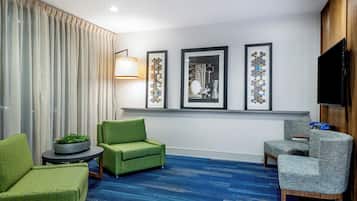 Una camera d'hotel con tappeto blu, poltrone rivestite in verde e grigio, un tavolino rotondo, una pianta in vaso e tre quadri appesi al muro.