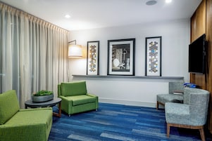 Una camera d'hotel con tappeto blu, poltrone rivestite in verde e grigio, un tavolino rotondo, una pianta in vaso e tre quadri appesi al muro.